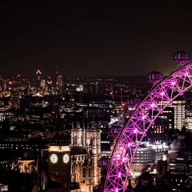 A section of the London Eye with pink lights glowing at night. Landmarks such as the Big Ben and Houses of Parliament can be seen illuminated in the background.