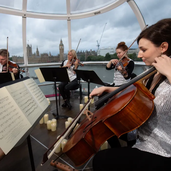A group of musicians surrounded by candlelight playing the violins and cello inside a London Eye Pod at dusk. Big Ben and the Houses of Parliament are visible in the distance.