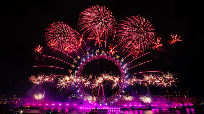The London Eye lit up in pink at night, with red, yellow and orange fireworks bursting around it during New Year's Eve celebrations.