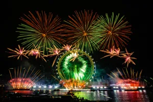 The London Eye lit up in green at night, with green, red, and orange fireworks bursting around it during New Year's Eve celebrations.