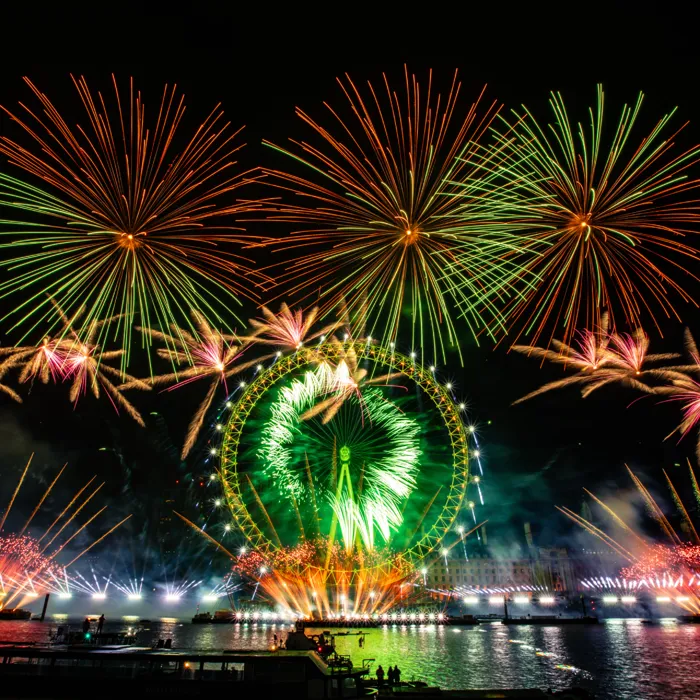 The London Eye lit up in green at night, with green, red, and orange fireworks bursting around it during New Year's Eve celebrations.