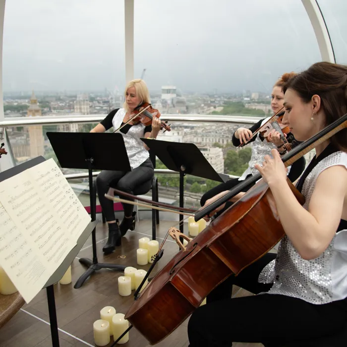 A group of musicians surrounded by candlelight playing the violins and cello inside a London Eye Pod at dusk.