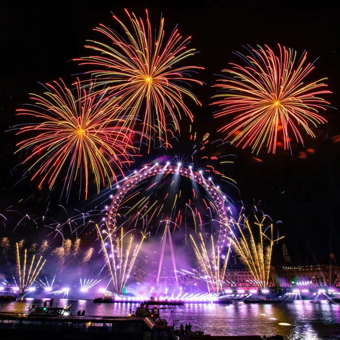 The London Eye lit up in violet at night, with multi-coloured fireworks bursting around it during New Year's Eve celebrations.