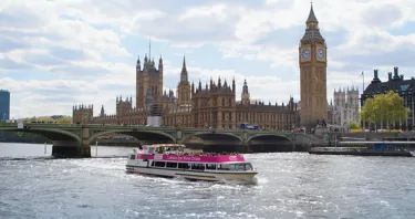 The London Eye River Cruise pictured after passing under Westminster Bridge on a partly sunny day. Big Ben and The Houses of Parliament are visible in the background.